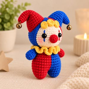 May include: A small crochet jester doll featuring a two-toned red and blue hat with gold jingle bells at the tips. The doll has a cream-colored face, black bead eyes, a red nose, a yellow ruffled collar, and a red and blue patterned body.