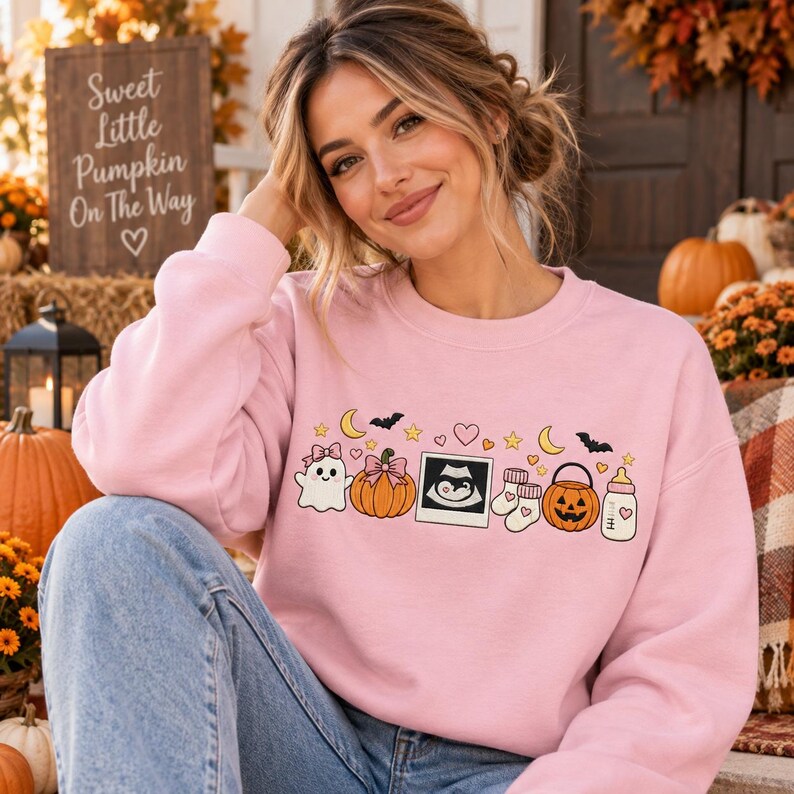 May include: A light pink crewneck sweatshirt featuring a graphic print of a ghost, a pumpkin with a bow, an ultrasound image, baby socks, a jack-o'-lantern bucket, and a baby bottle, surrounded by small stars, hearts, moons, and bats.