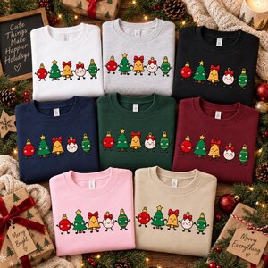 May include: A collection of eight crewneck sweatshirts in white, heather gray, black, navy, dark green, maroon, pink, and beige. Each features a graphic of five cartoon Christmas ornaments holding hands, including a tree, bell, and baubles.