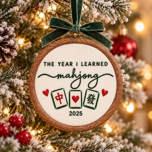 May include: A round wooden embroidery hoop ornament featuring cream fabric with green embroidered text that reads THE YEAR I LEARNED mahjong 2025. The design includes three mahjong tiles with red and green details, two red hearts, and a dark green velvet ribbon bow.