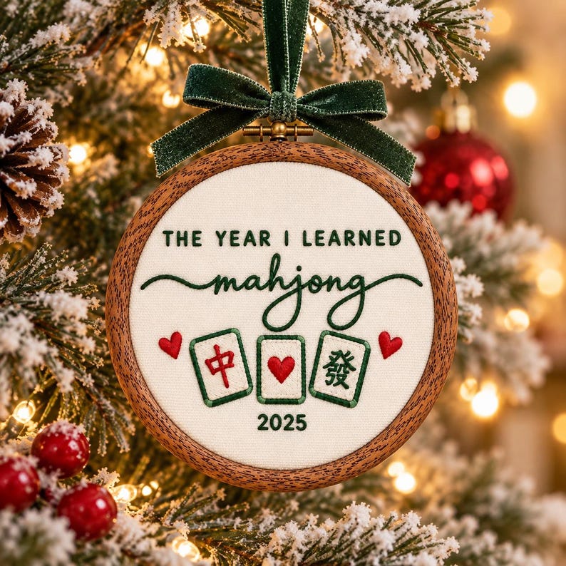 May include: A round wooden embroidery hoop ornament featuring cream fabric with green embroidered text that reads THE YEAR I LEARNED mahjong 2025. The design includes three mahjong tiles with red and green details, two red hearts, and a dark green velvet ribbon bow.