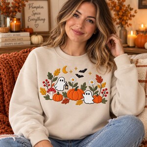 May include: A beige crewneck sweatshirt featuring a central embroidered design of two white ghosts, orange pumpkins, red mushrooms, yellow autumn leaves, white daisies, a yellow crescent moon, and two black bats against a neutral background.
