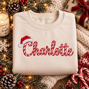 May include: A beige crewneck sweatshirt featuring the name Charlotte embroidered in a red and white candy cane striped pattern. A small red and white Santa hat is positioned above the letter C, completing the festive design on the front.