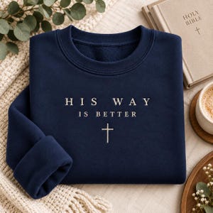 May include: A navy blue crewneck sweatshirt featuring the embroidered white text "HIS WAY IS BETTER" centered on the chest, positioned above a small, simple white cross symbol. The garment has long sleeves and a ribbed collar, cuffs, and hem.