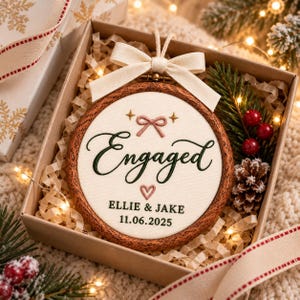May include: A round wooden embroidery hoop ornament featuring cream fabric with the embroidered text Engaged, a pink bow, a small heart, and the names Ellie & Jake with the date 11.06.2025. The hoop is topped with a cream ribbon bow.