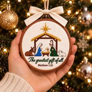 May include: A circular wooden embroidery hoop ornament featuring a nativity scene with Mary, Joseph, and the infant Jesus in a manger under a gold star. The text reads The greatest gift of all and Matthew 1:21. A cream ribbon bow is at the top.