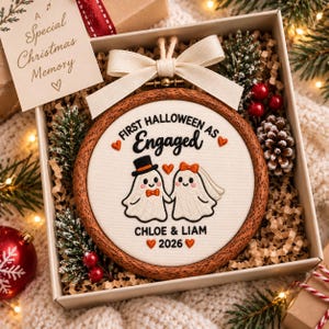 May include: A round wooden embroidery hoop ornament featuring white fabric with stitched text reading FIRST HALLOWEEN AS Engaged, CHLOE & LIAM 2026, and two ghost figures wearing a top hat and bow tie, and a veil and bow, accented with orange hearts.