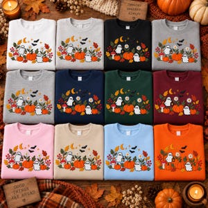 May include: A collection of twelve crewneck sweatshirts in various colors, each featuring a central graphic of ghosts, pumpkins, mushrooms, autumn leaves, flowers, stars, and bats. The sweatshirts are arranged in a grid on a wooden surface.