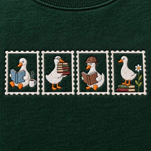May include: A dark green sweatshirt featuring four embroidered stamp-shaped designs. Each frame depicts a white duck with orange feet and a beak, engaged in various activities including reading books, holding a stack of books, and standing near a flower.