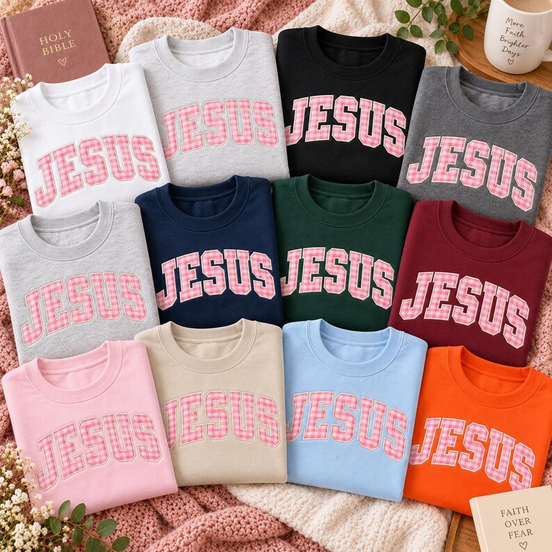May include: A collection of twelve crewneck sweatshirts in various colors including white, gray, black, navy, dark green, maroon, pink, beige, light blue, and orange. Each sweatshirt features the word JESUS in pink and white gingham letters.