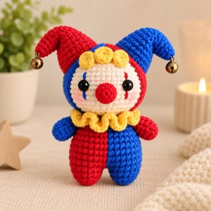 May include: A crocheted clown doll featuring a two-toned red and blue hat with small metallic bells at the tips. The doll has a cream-colored face, a red nose, black eyes, blue tear-drop markings, and a yellow ruffled collar around its neck.