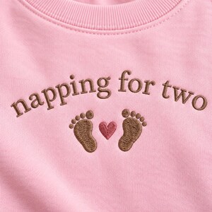 May include: A light pink sweatshirt featuring brown embroidered text that reads napping for two, positioned above two brown embroidered baby footprints and a small pink embroidered heart centered between the footprints on the chest area.