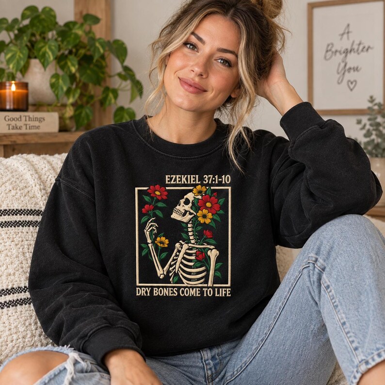 May include: A black crewneck sweatshirt featuring a graphic print of a skeleton surrounded by red and yellow flowers. The text above the graphic reads EZEKIEL 37:1-10 and the text below reads DRY BONES COME TO LIFE in a cream-colored font.