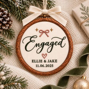May include: A round wooden embroidery hoop ornament featuring cream fabric with black embroidered text reading Engaged, Ellie & Jake, and 11.06.2025. The design includes a pink embroidered bow, a small heart, and two gold stars, topped with a cream ribbon.