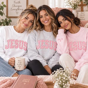 May include: Three crewneck sweatshirts in white, heather gray, and light pink, each featuring the word JESUS in pink and white gingham block letters. The pink sweatshirt includes the text John 3:16 and a small heart embroidered on the sleeve.