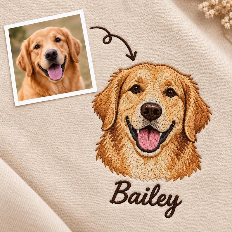 May include: A beige fabric sweatshirt featuring a detailed golden retriever embroidery portrait. Above the dog's head is a dark brown embroidered arrow pointing to a square photograph of the same dog. The name Bailey is embroidered in dark brown script below.