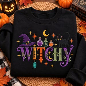 May include: A black sweatshirt featuring a colorful graphic with the text WITCHY in large letters. The design includes a purple witch hat, three small potion bottles, a black cat, a yellow crescent moon, orange stars, and a hanging crystal.