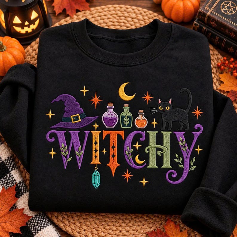 May include: A black sweatshirt featuring a colorful graphic with the text WITCHY in large letters. The design includes a purple witch hat, three small potion bottles, a black cat, a yellow crescent moon, orange stars, and a hanging crystal.