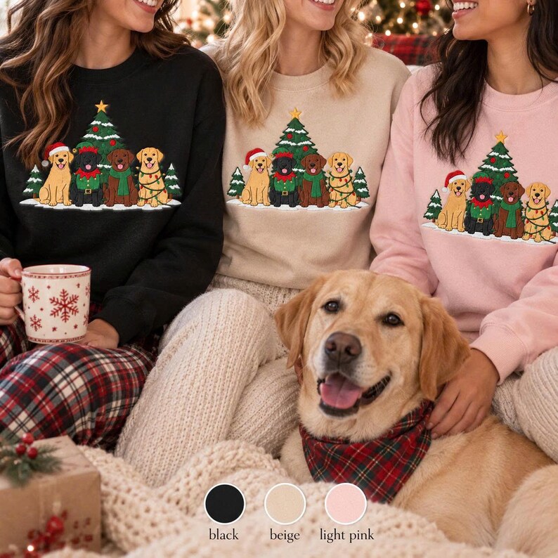 May include: Three crewneck sweatshirts in black, beige, and light pink, each featuring a graphic print of four dogs sitting in front of a decorated Christmas tree. The dogs are wearing festive accessories like Santa hats and scarves.