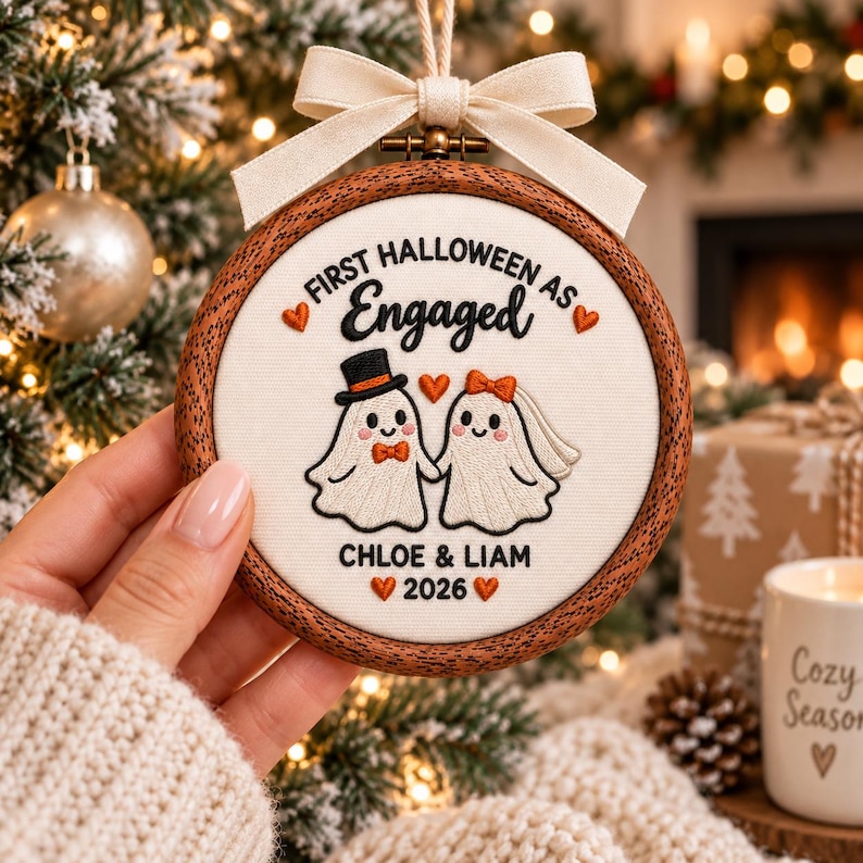 May include: A round wooden embroidery hoop ornament featuring white fabric with embroidered text First Halloween as Engaged, Chloe & Liam, and 2026. The design includes two ghost figures, one with a top hat and one with a veil, accented by orange hearts and a cream-colored ribbon bow at the top.