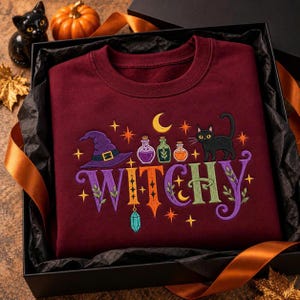 May include: A maroon crewneck sweatshirt featuring an embroidered design with the word WITCHY in colorful letters, a purple witch hat, three small potion bottles, a black cat, a yellow crescent moon, and several small orange stars.