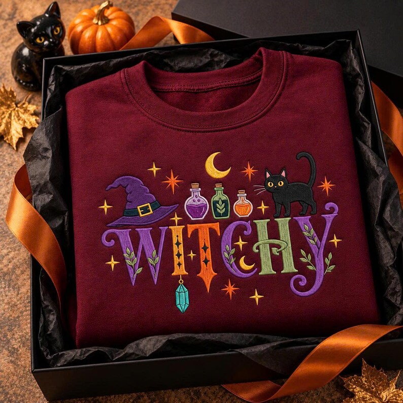 May include: A maroon crewneck sweatshirt featuring an embroidered design with the word WITCHY in colorful letters, a purple witch hat, three small potion bottles, a black cat, a yellow crescent moon, and several small orange stars.