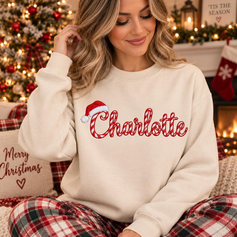 May include: A beige crewneck sweatshirt featuring the name Charlotte embroidered in a red and white candy cane striped font. A small red and white Santa hat is positioned above the letter C. The sweatshirt has long sleeves and a ribbed collar.