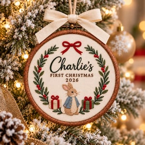 May include: A round wooden embroidery hoop ornament featuring a cream fabric centre with a green holly wreath, a red bow, and a brown rabbit wearing a blue jacket. The text reads Charlie's FIRST CHRISTMAS 2026, flanked by two gift boxes.