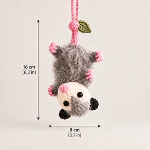 May include: A crocheted opossum hanging by its tail, featuring a fuzzy gray body, a white face with black eyes, pink nose and feet, and a green leaf accent. The hanging ornament measures 6.3 inches in height and 3.1 inches in width.