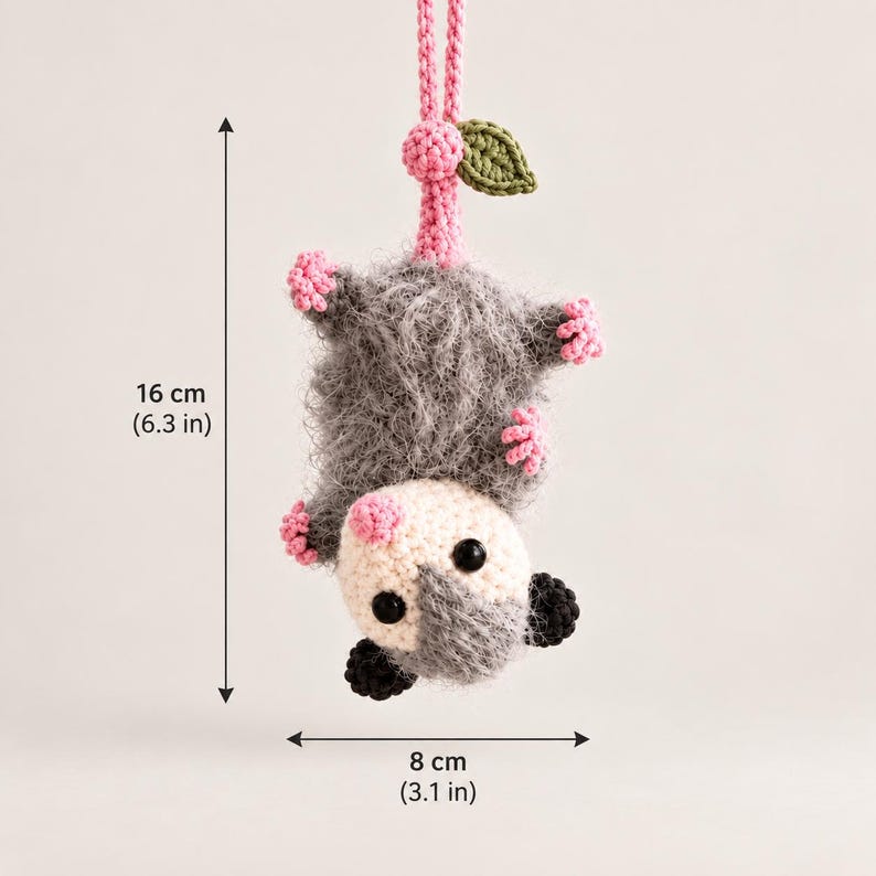 May include: A crocheted opossum hanging by its tail, featuring a fuzzy gray body, a white face with black eyes, pink nose and feet, and a green leaf accent. The hanging ornament measures 6.3 inches in height and 3.1 inches in width.