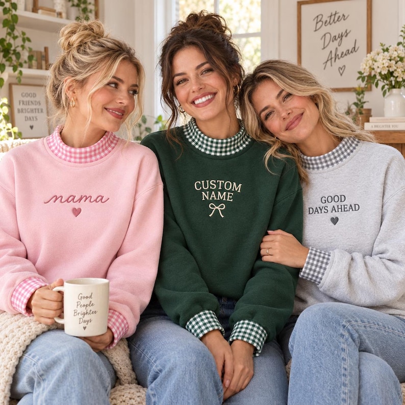 May include: Three long-sleeve sweatshirts with mock neck collars and cuffs featuring a gingham pattern. The pink sweatshirt has the text "nama" and a heart, the green one says "CUSTOM NAME" with a bow, and the gray one says "GOOD DAYS AHEAD".