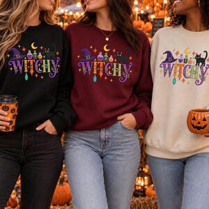 May include: Three crewneck sweatshirts in black, maroon, and beige, each featuring a graphic print with the text WITCHY, a purple witch hat, colorful potion bottles, a black cat, a yellow crescent moon, and various stars and sparkles.