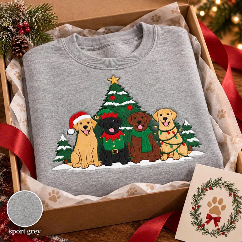 May include: A heather gray crewneck sweatshirt featuring a graphic of four Labrador dogs in front of a Christmas tree. The dogs include a yellow Labrador in a Santa hat, a black Labrador in an elf outfit, a chocolate Labrador in a green scarf, and a yellow Labrador wrapped in colorful Christmas lights.