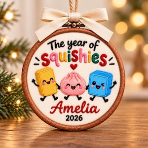 May include: A circular wooden embroidery hoop ornament featuring white fabric with the embroidered text The year of squishies, a small red heart, and three smiling cartoon characters in yellow, pink, and blue. The name Amelia and 2026 are stitched below, topped with a cream ribbon.