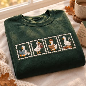 May include: A dark green crewneck sweatshirt featuring an embroidered design of four white postage stamp frames. Each frame contains a white duck engaged in reading or carrying books, with one duck wearing a hat and another near a cup of coffee.