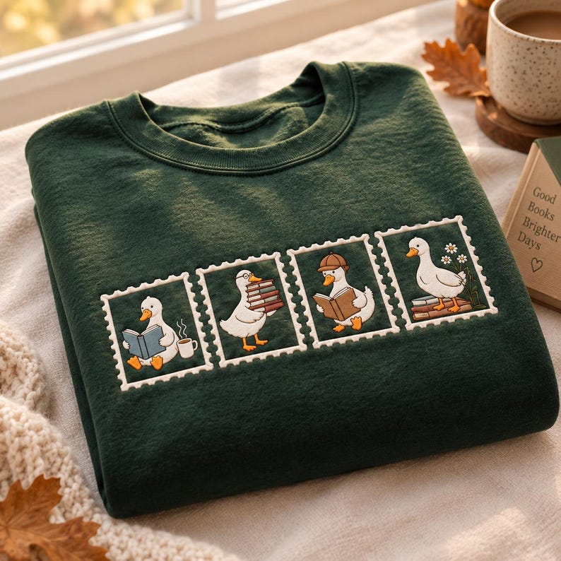May include: A dark green crewneck sweatshirt featuring an embroidered design of four white postage stamp frames. Each frame contains a white duck engaged in reading or carrying books, with one duck wearing a hat and another near a cup of coffee.