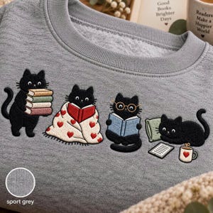 May include: A sport grey crewneck sweatshirt featuring an embroidered design of four black cats. One cat holds a stack of books, one is wrapped in a heart-patterned blanket reading, one wears glasses while reading, and one lies by a book and mug.