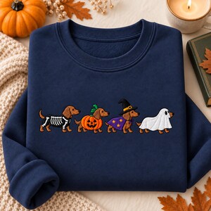 May include: A navy blue crewneck sweatshirt featuring an embroidered graphic of four brown dachshunds in Halloween costumes. The dogs are dressed as a skeleton, a jack-o'-lantern, a witch with a purple cape and hat, and a white ghost sheet.