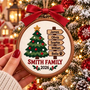 May include: A round wooden embroidery hoop ornament featuring a stitched green Christmas tree with a gold star, a signpost with the names James, Olivia, Sophia, William, and Bella, and the text Smith Family 2026 with a red ribbon bow on top.