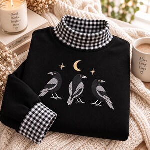 May include: A black long-sleeve sweatshirt featuring a mock neck collar and sleeve cuffs in a black and white gingham pattern. The front displays an embroidered design of three black crows positioned beneath a gold crescent moon and two stars.