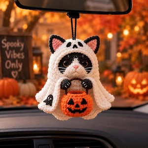 May include: A crocheted cat ornament wearing a white ghost costume with black ears and a small black ghost detail. The cat holds a bright orange crocheted pumpkin bucket with a jack-o'-lantern face. It hangs from a black cord for display.