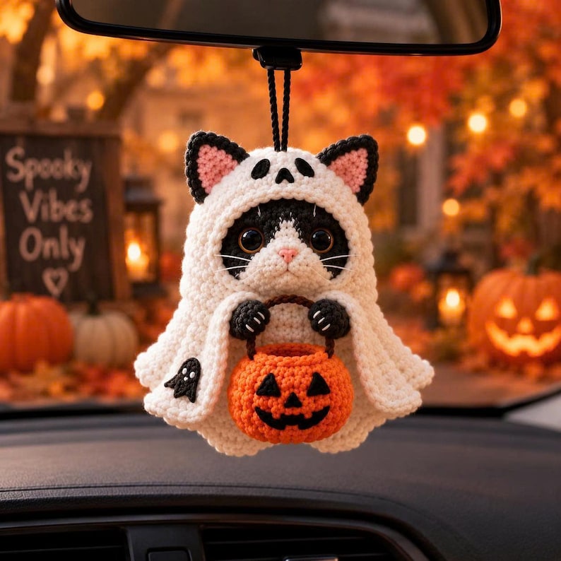 May include: A crocheted cat ornament wearing a white ghost costume with black ears and a small black ghost detail. The cat holds a bright orange crocheted pumpkin bucket with a jack-o'-lantern face. It hangs from a black cord for display.