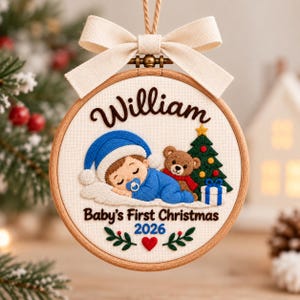 May include: A round wooden embroidery hoop ornament featuring a cream fabric background. It displays the name William, a sleeping baby in a blue hat and outfit with a pacifier, a teddy bear, a Christmas tree, and the text Baby's First Christmas 2026.