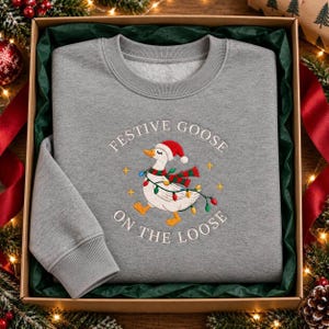 May include: A heather gray crewneck sweatshirt featuring an embroidered graphic of a white goose wearing a red and white Santa hat and a red and green striped scarf. The goose is wrapped in colorful Christmas lights with the text Festive Goose On The Loose.