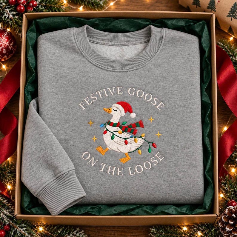 May include: A heather gray crewneck sweatshirt featuring an embroidered graphic of a white goose wearing a red and white Santa hat and a red and green striped scarf. The goose is wrapped in colorful Christmas lights with the text Festive Goose On The Loose.