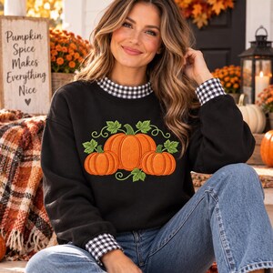 May include: A black long-sleeve sweatshirt featuring an embroidered graphic of three orange pumpkins with green leaves and vines. The garment has a black and white gingham check pattern on the inner collar and sleeve cuffs, paired with blue jeans.
