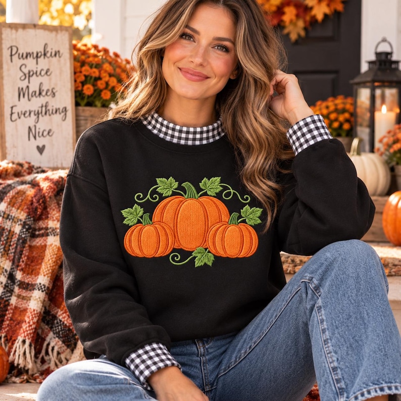 May include: A black long-sleeve sweatshirt featuring an embroidered graphic of three orange pumpkins with green leaves and vines. The garment has a black and white gingham check pattern on the inner collar and sleeve cuffs, paired with blue jeans.