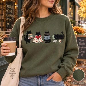 May include: An olive green crewneck sweatshirt featuring a graphic of four black cats engaged in reading activities. One cat holds a stack of books, another is wrapped in a heart-patterned blanket, one wears glasses while reading, and one lies by a mug.