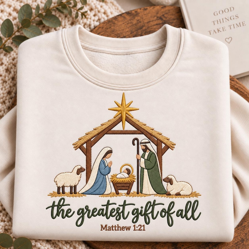 May include: A cream-colored crewneck sweatshirt featuring an embroidered nativity scene with a gold star, a manger, and figures in blue and green robes. Below the scene, the text reads "the greatest gift of all" and "Matthew 1:21" in dark green.