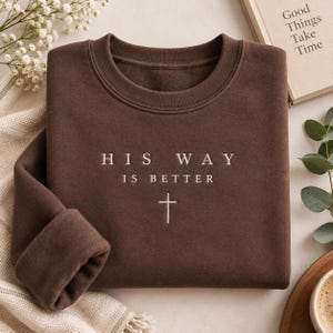 May include: A dark brown crewneck sweatshirt featuring the embroidered text "HIS WAY IS BETTER" in a light cream color, positioned above a small, simple cross symbol centered on the chest of the garment.